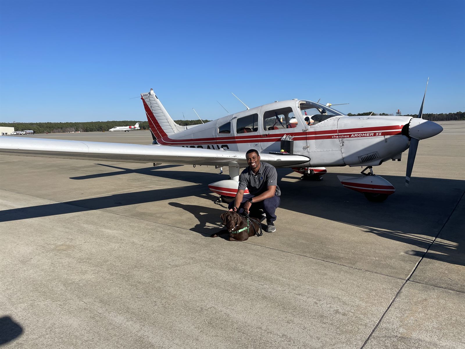 Cam kneeling with a chocolate lab in front of a Piper Archer